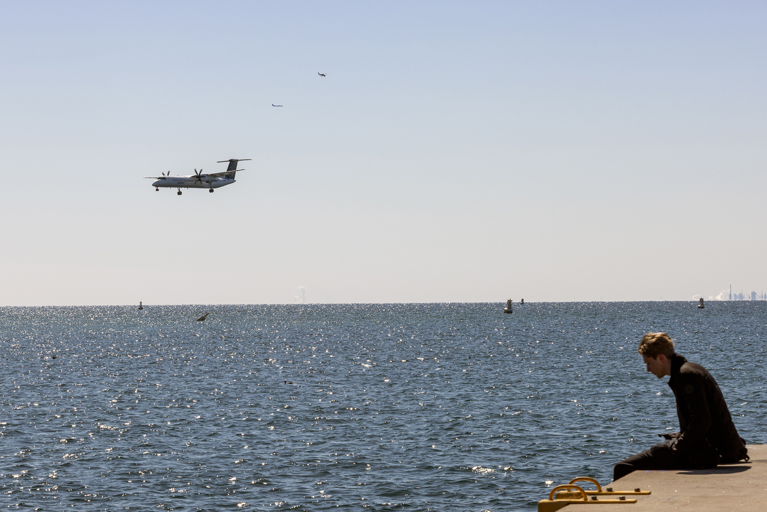 Porter Airlines turboprop approaches Billy Bishop Airport from the west, while a man sits on the pier of the Western Channel that links Toronto Harbour and Lake Ontario. 