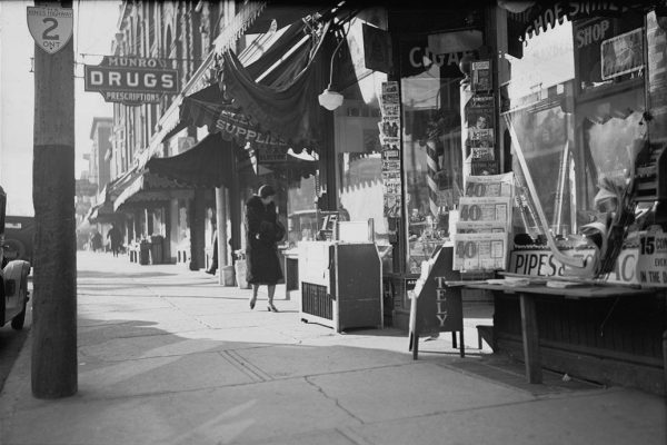 Black and white photograph of series of storefronts, with woman looking into one of them